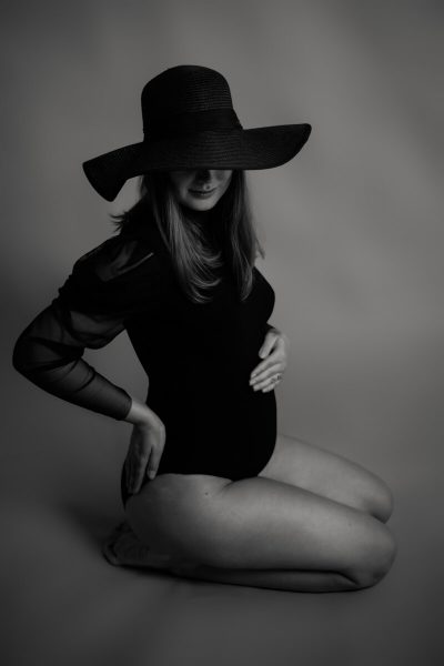 die Schwangere kniet am Boden und hat einen schwarzen Body an und einen Hut auf in schwarz/weiß